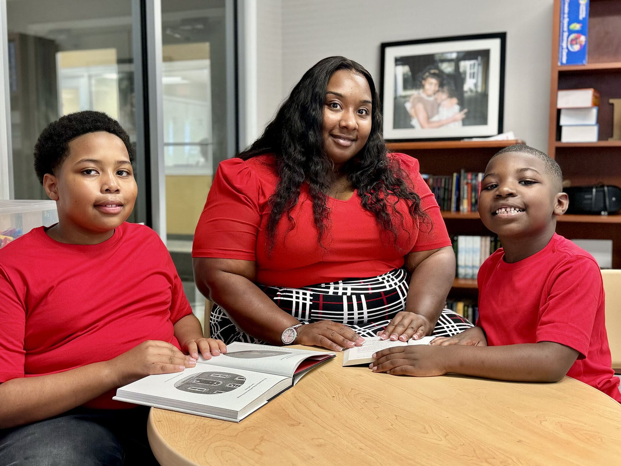 A mother and two sons in red shirts reading books at a table.